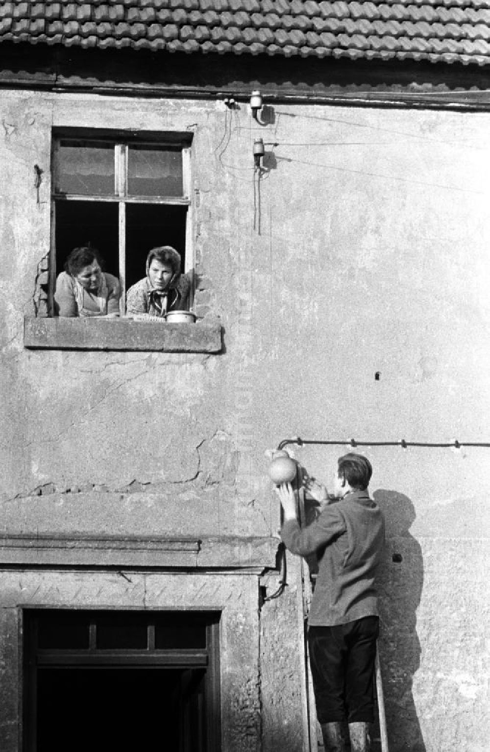 GDR photo archive: Leipzig - Ein Mann steht an einer Hauswand auf einer Leiter und repariert eine Lampe, zwei Frauen schauen ihm aus dem Fenster heraus dabei zu.