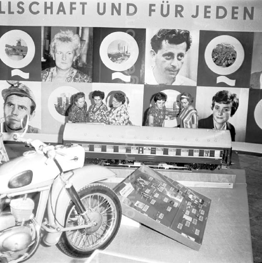 GDR image archive: Berlin - Ausstellungsbesucher scheuen sich Exponate an, im Hintergrund großes Plakat mit Gesichtern. Vorne steht ein Motarad der Marke MZ, dahinter ein Model vom Zuganhänger. Anläßlich zum 7