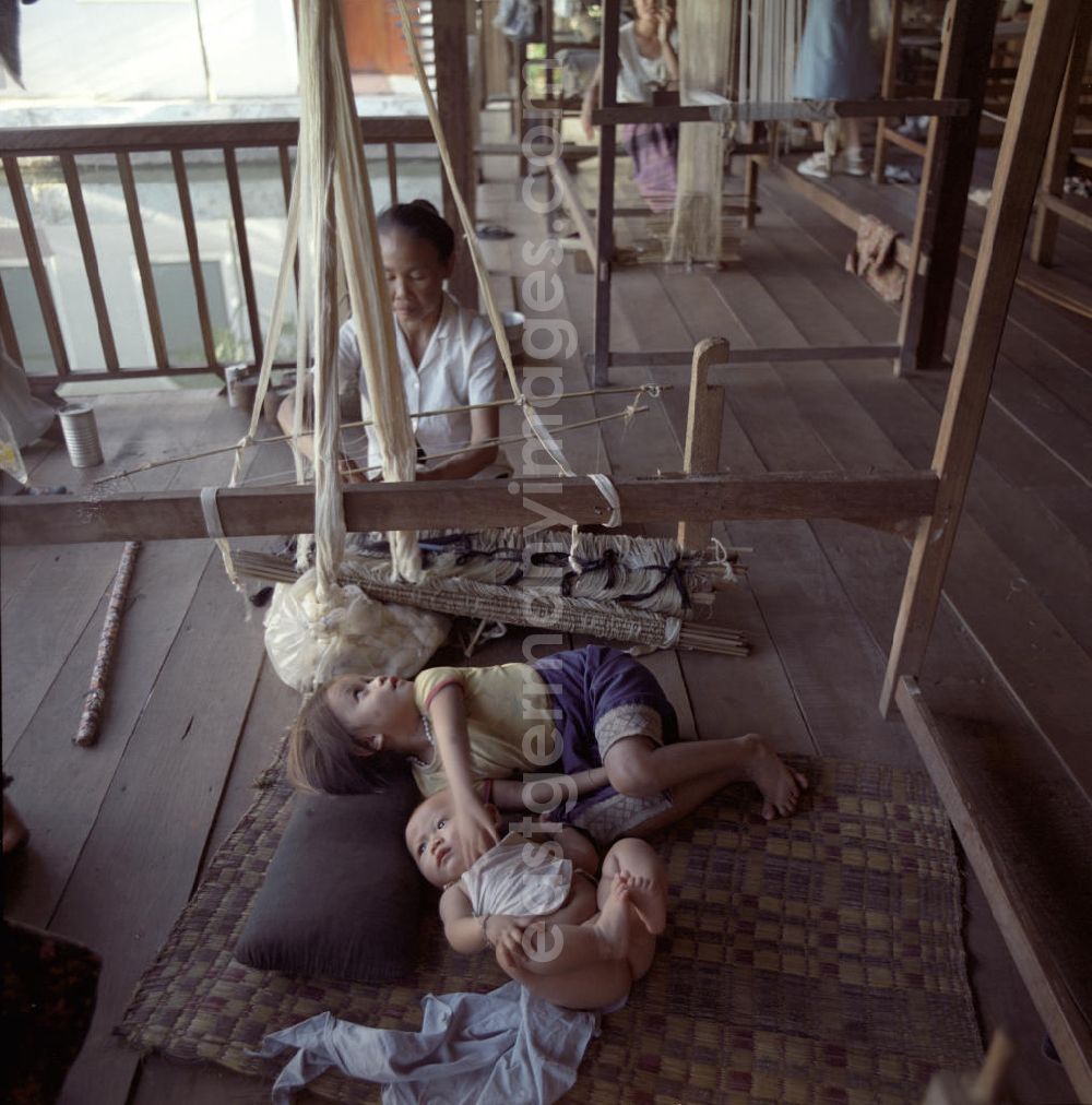 GDR picture archive: Vientiane - Eine Weberin bei ihrer Arbeit, während die Kinder daneben auf einer Matte liegen in einer Weberei in Vientiane, der Hauptstadt der Demokratischen Volksrepublik Laos.