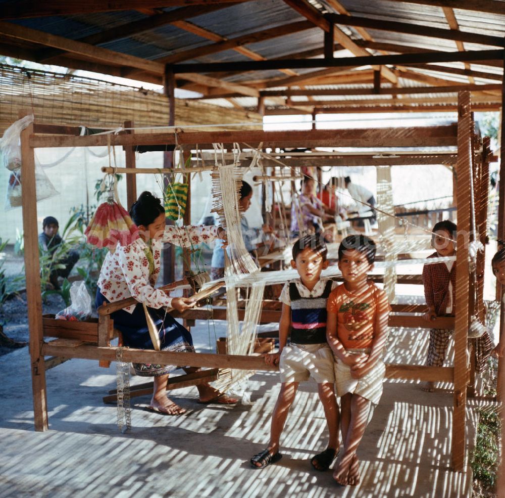 GDR picture archive: Vientiane - Frauen bei ihrer Arbeit, während die Kinder daneben spielen in einer Weberei in Vientiane, der Hauptstadt der Demokratischen Volksrepublik Laos.