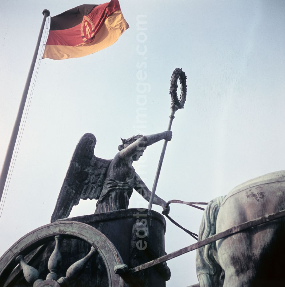 Berlin: GDR flag - Tourist attraction and landmark Brandenburg Gate on Pariser Platz in the Mitte district of East Berlin in the area of the former GDR, German Democratic Republic. The statue group of the quadriga created by Johann Gottfried Schadow on the gate built by Carl Gotthard Langhans in the early classicist style is well known