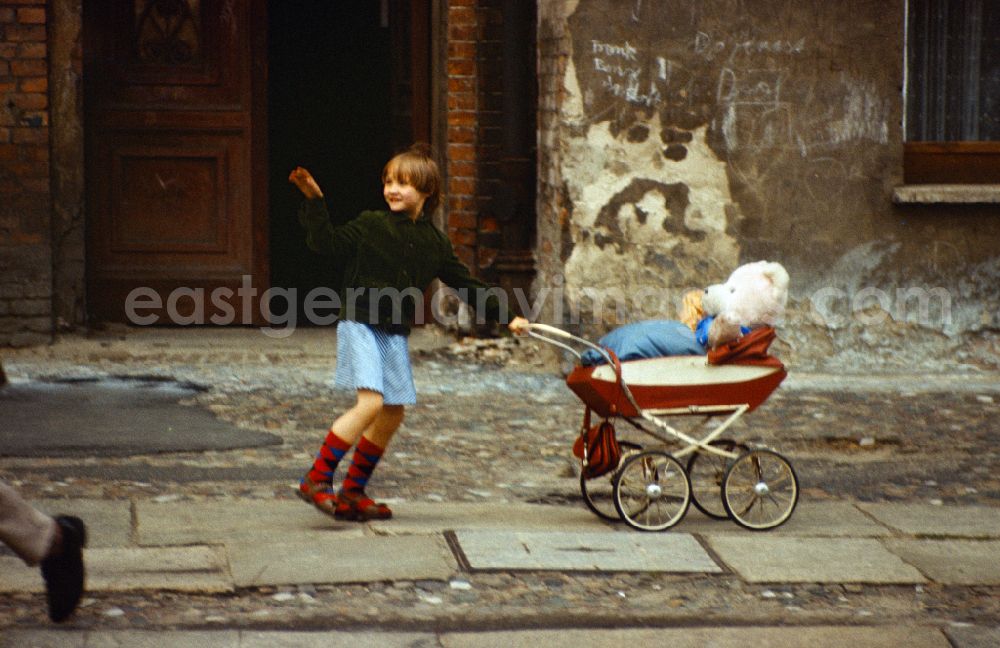 GDR image archive: Berlin - Girl pushes her teddy bear in a doll's pram on a sidewalk in East Berlin on the territory of the former GDR, German Democratic Republic