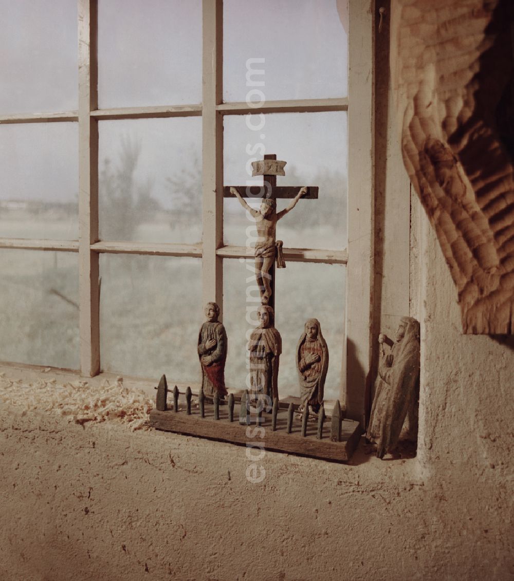 GDR image archive: Räckelwitz - Scene from the film and television production Portrait of a Center Wooden crucifixes in the workshop of the wood restorer Benedikt Dirlich on the main street in Raeckelwitz, Saxony in the area of the former GDR, German Democratic Republic. In front of a window stands a group of wooden figures and a cross with Corpus Christie