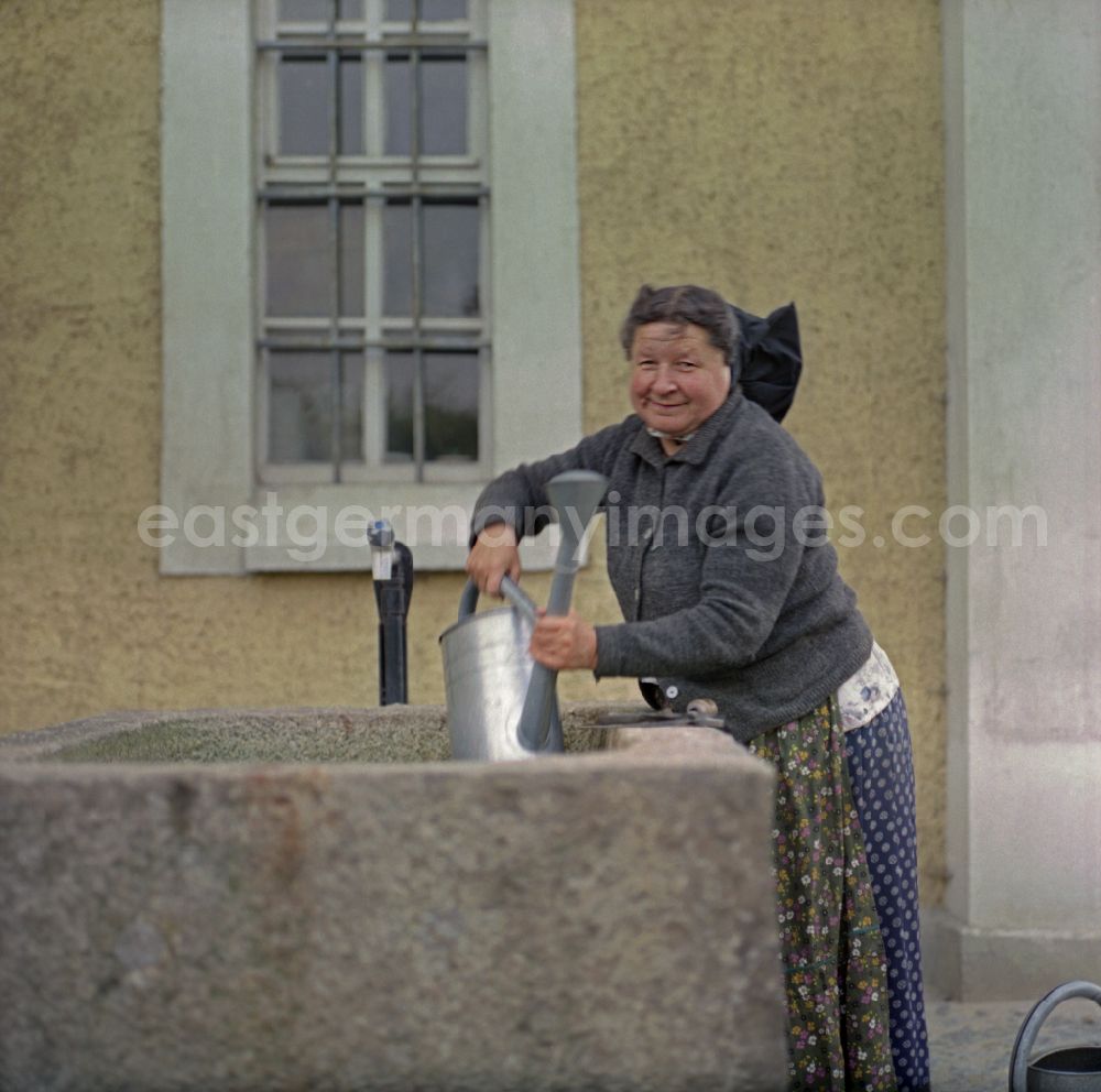 GDR image archive: Ralbitz-Rosenthal - Scene from the film and television production Portrait of a Center on the main street in Ralbitz-Rosenthal, Saxony in the area of the former GDR, German Democratic Republic. A brick water basin. A Sorbian woman in everyday dress lifts a metal watering can out of the water and looks into the camera