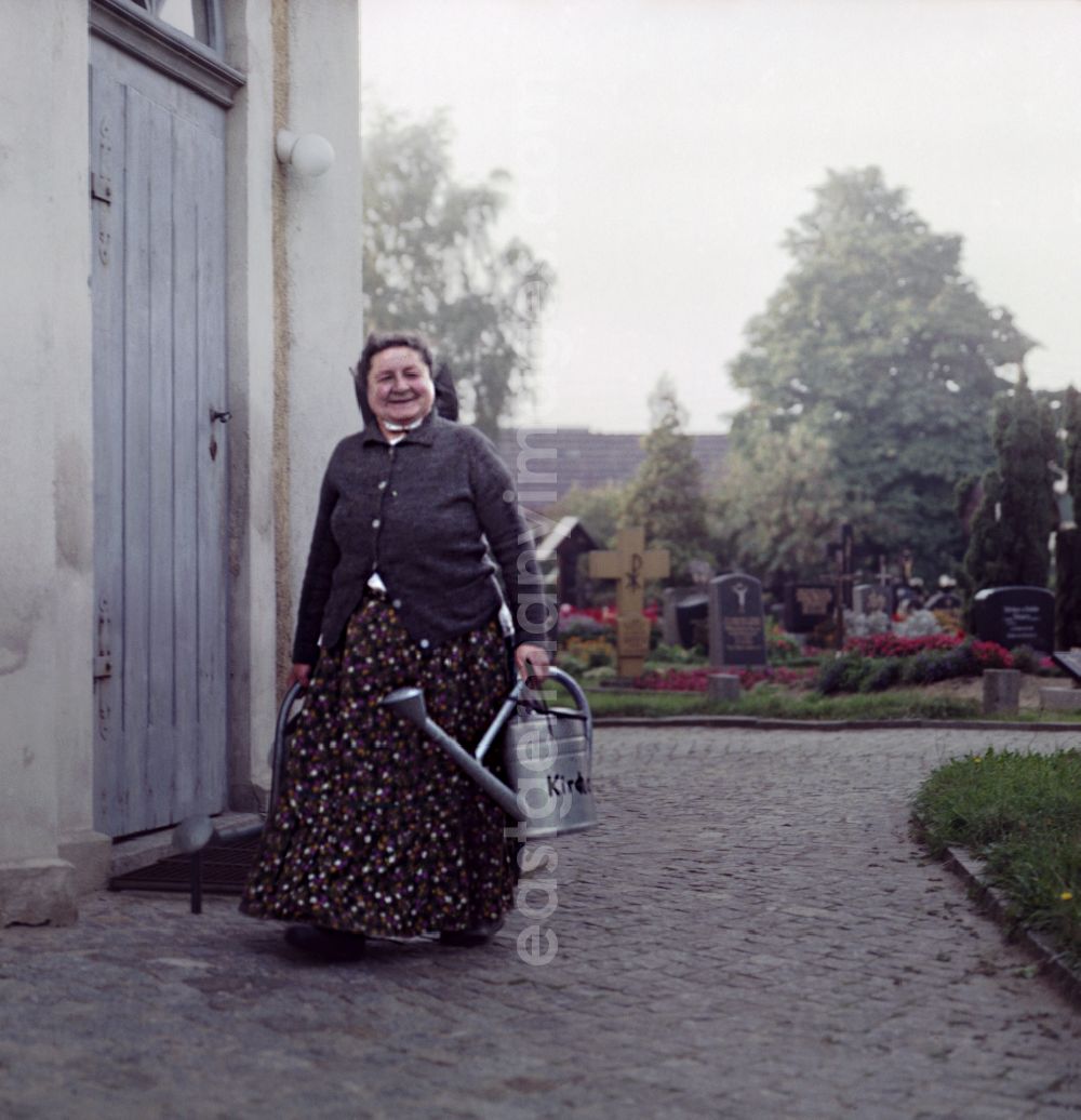 GDR photo archive: Ralbitz-Rosenthal - A Sorbian woman in everyday costume carries a metal watering can as a scene from the film and television production Portrait of a Center Sorbian cemetery in Ralbitz-Rosenthal, Saxony in the area of the former GDR, German Democratic Republic