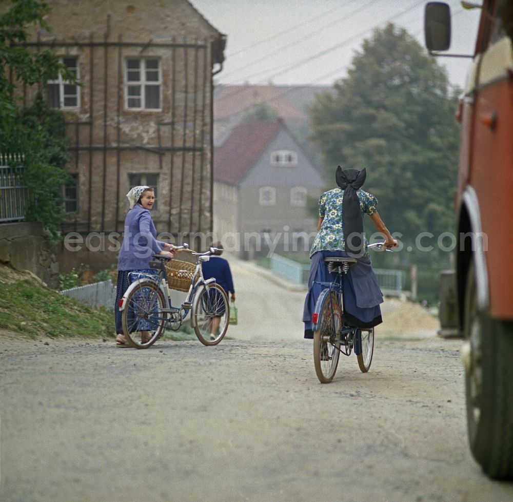 GDR photo archive: Räckelwitz - Two female cyclists in traditional peasant clothing meet on a village street as a scene from the film and television production Portrait of a Center on the main street in Raeckelwitz, Saxony in the area of the former GDR, German Democratic Republic