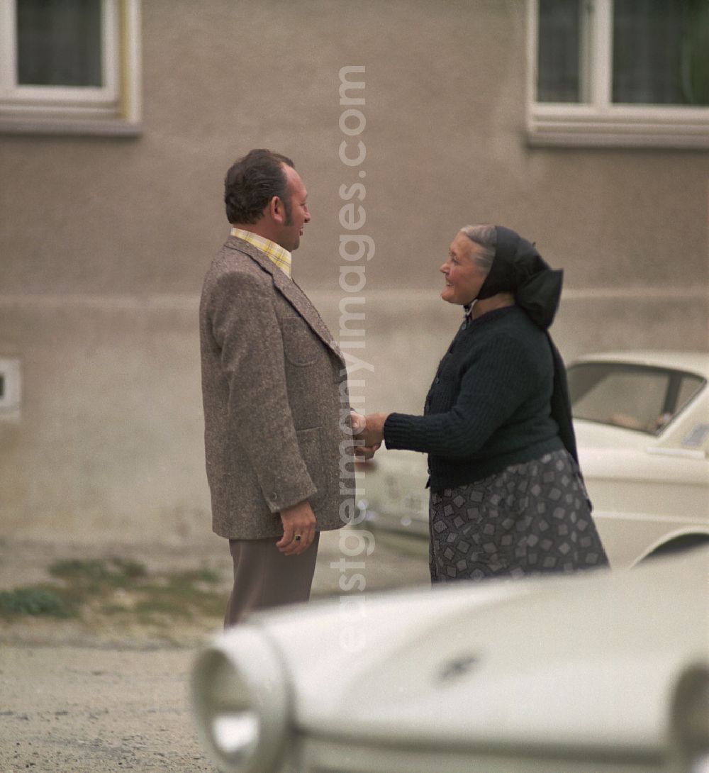 GDR picture archive: Räckelwitz - On a village street, a man and an elderly woman in Sorbian everyday costume stand between two Trabant cars and have an animated conversation as a scene from the film and television production Portrait of a Center on the main street in Raeckelwitz, Saxony in the area of the former GDR, German Democratic Republic