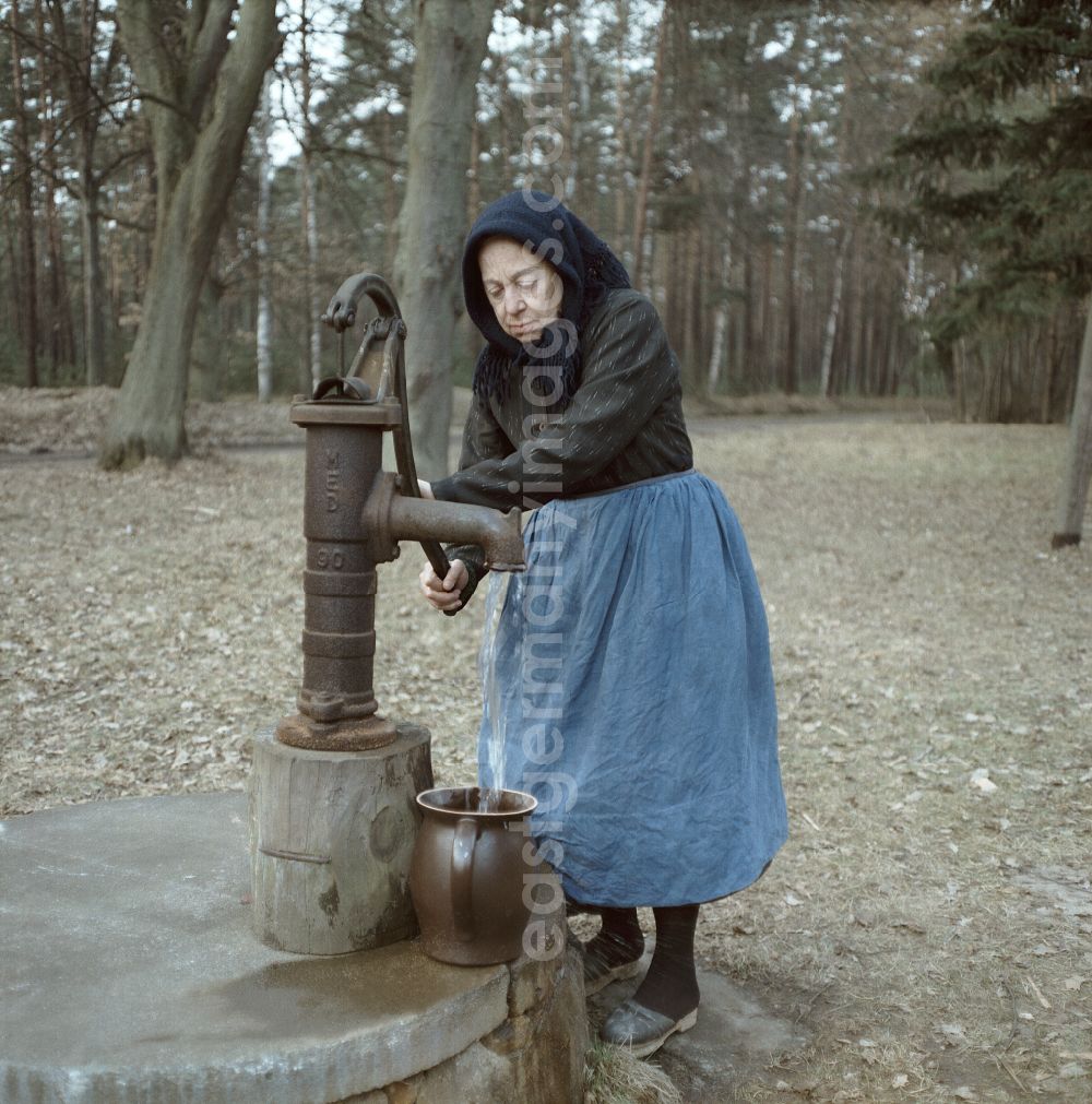 GDR picture archive: Ortschaft Heide - Scene from the film and television production Rublak - The Legend of the Surveyed Land in Heide, Saxony, in the area of the former GDR, German Democratic Republic. An old farmer's wife in everyday dress stands at the edge of a forest at a hand pump. Water flows into a clay jug. The actress Doris Thalmer as a Sorbian farmer's wife