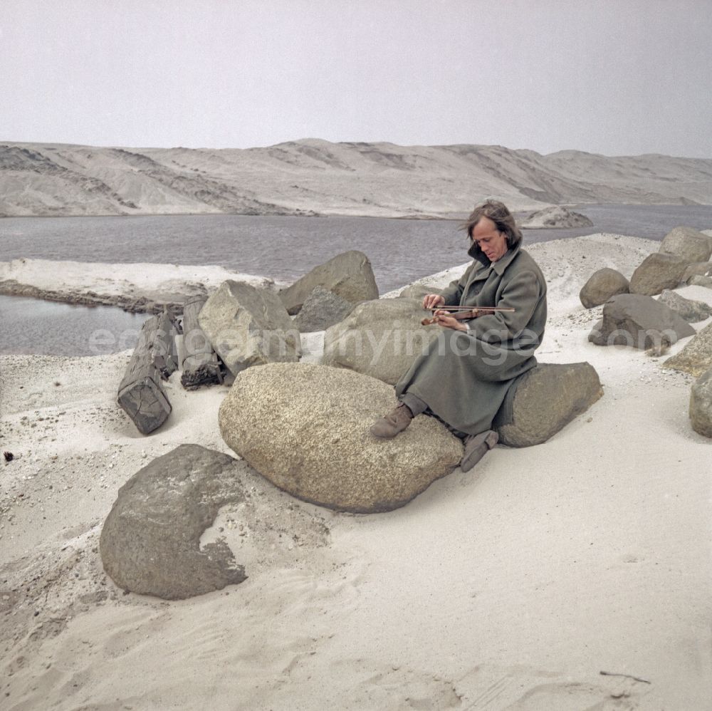 GDR photo archive: Weißwasser/Oberlausitz - Scene from the film and television production Rublak - The Legend of the Surveyed Land in Weisswasser/Oberlausitz, Saxony in the area of the former GDR, German Democratic Republic. The actor Christian Grashof sits playing the violin on a very large boulder in the middle of a sandy plain in a waste landscape of a brown coal open-cast mine