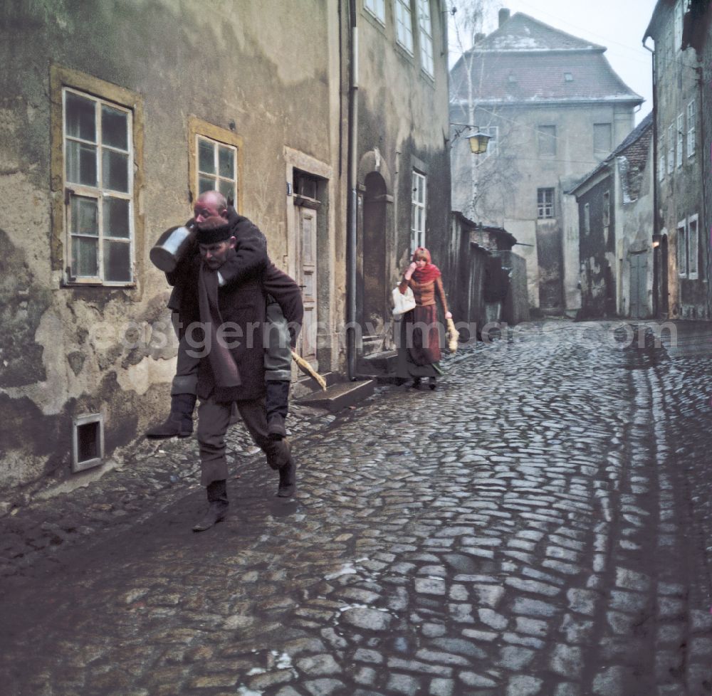 Bautzen: Scene photo for the film: Continent Hope An evening street with cobblestones. A man with a straw broom (Mericin Slowdenk) carries a bald man (Axel Reinsahen) piggyback, who is drinking from a clay jug. A woman in a headscarf (Majka Ulbrichec) runs after them in the background