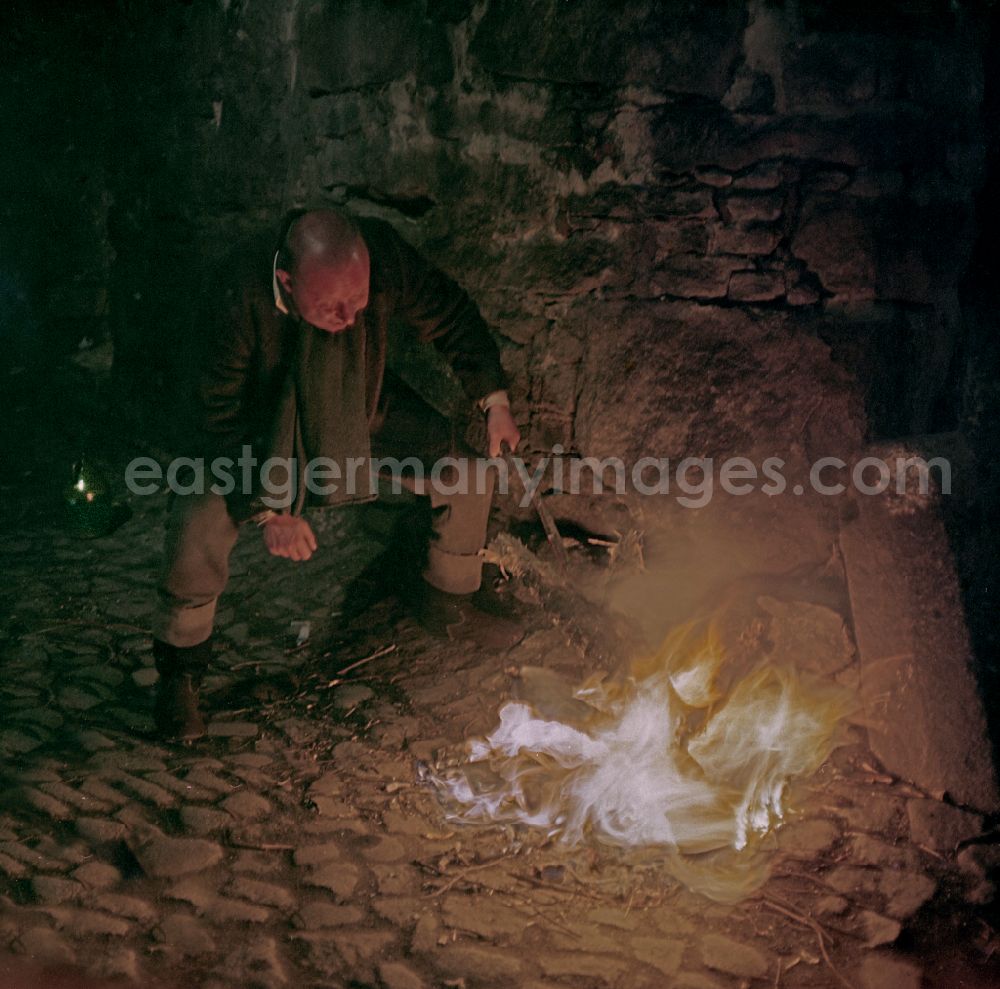 GDR photo archive: Bautzen - Scene photo for the film: Continent of Hope A small wood fire burns on the wall of a cobblestone street. A man (Axel Reinshagen) crouches in front of it and pokes the fire with a stick