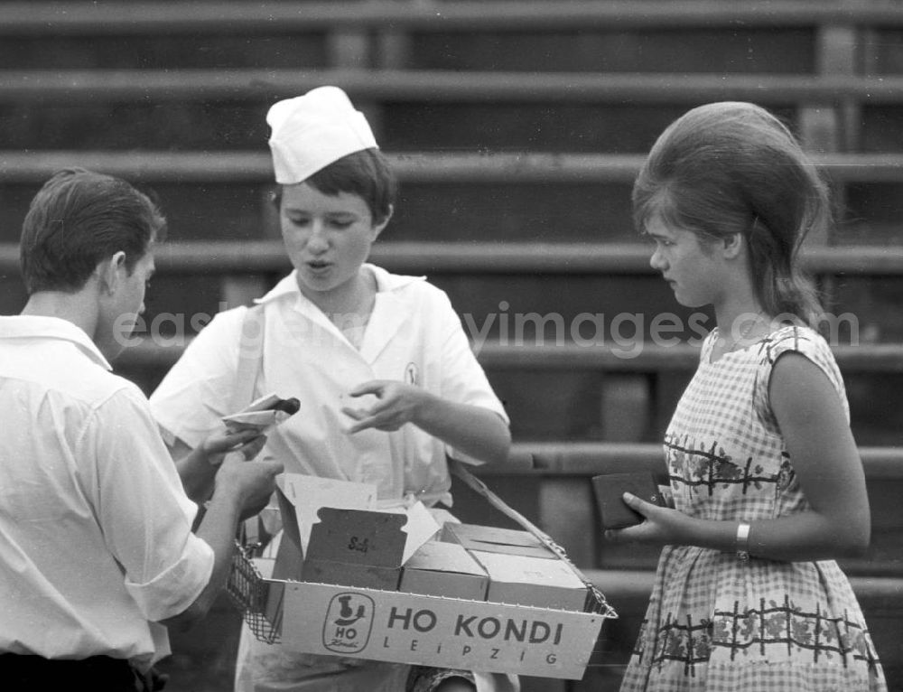 GDR picture archive: Leipzig - Eine Verkäuferin der HO Kondi - mit Bauchladen - verkauft Süßigkeiten auf dem Turnfest. Neben ihr ein Mädchen mit Toupet-Frisur und einem Kleid im Stil der 196