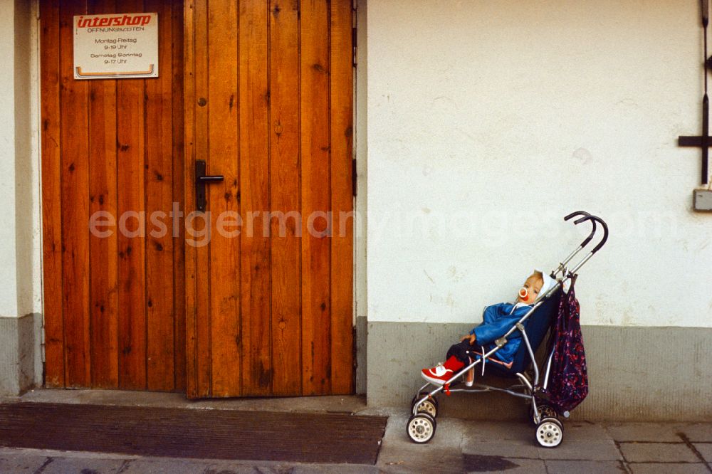 GDR photo archive: Berlin - Child sits in a buggy in front of an Intershop entrance in East Berlin in the territory of the former GDR, German Democratic Republic