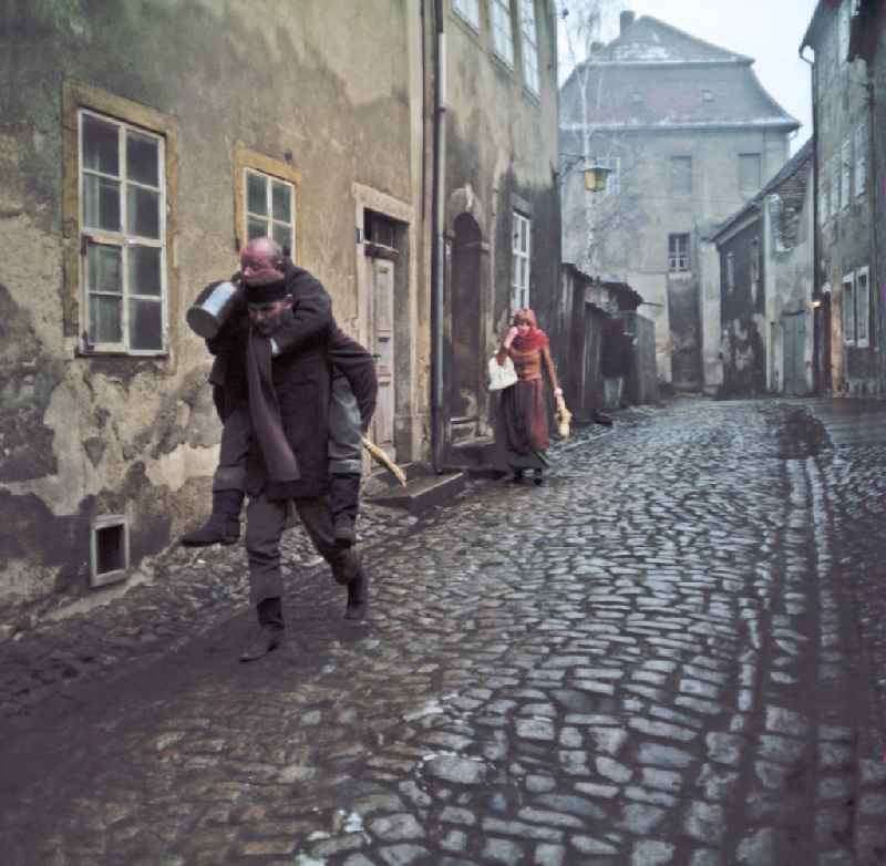 Scene photo for the film: 'Continent Hope' An evening street with cobblestones. A man with a straw broom (Mericin Slowdenk) carries a bald man (Axel Reinsahen) piggyback, who is drinking from a clay jug. A woman in a headscarf (Majka Ulbrichec) runs after them in the background