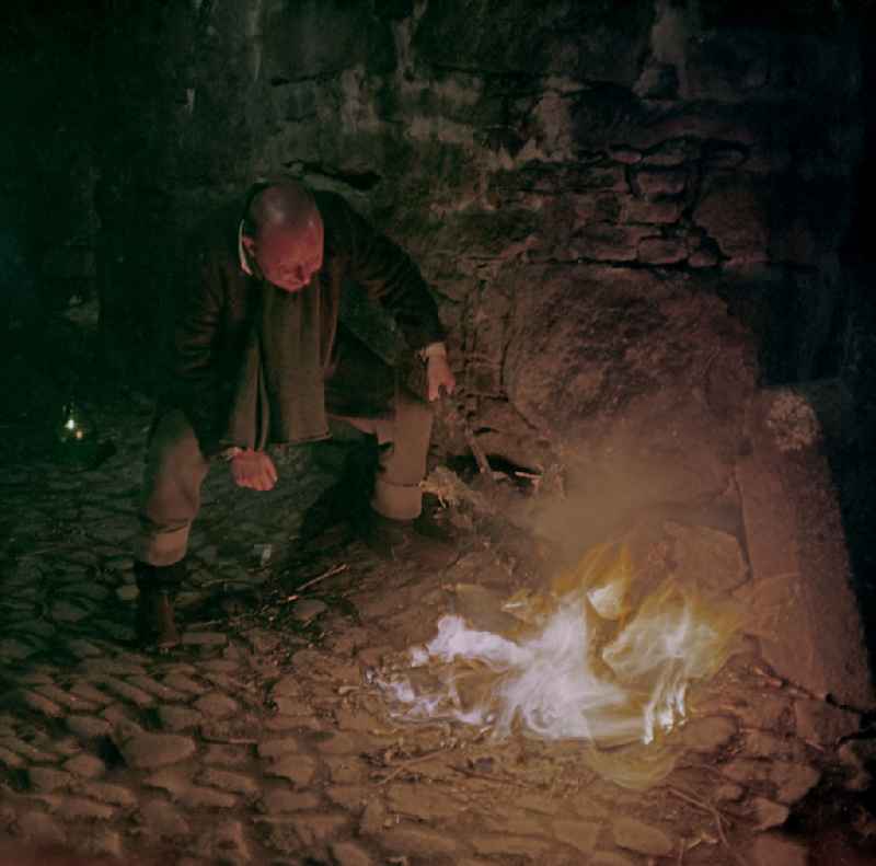 Scene photo for the film: 'Continent of Hope' A small wood fire burns on the wall of a cobblestone street. A man (Axel Reinshagen) crouches in front of it and pokes the fire with a stick