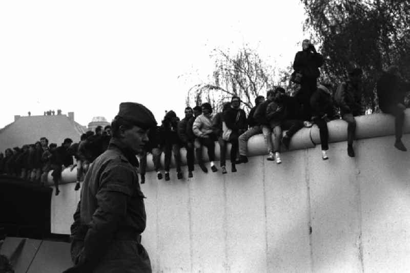 Soldaten der DDR stehen in einer Reihe vor der Berliner-Mauer am Potzdamer Platz, auf der Mauer sitzen und stehen Menschen.