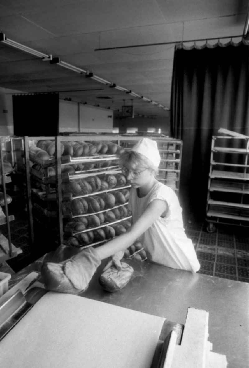 Eine Arbeiterin einer Bäckerei in Berlin-Heinersdorf nimmt die fertig gebackenen und noch heißen Brote / Brotlaibe vom Fließband. Im Hintergrund sind Mischbrote im Regal gestapelt.