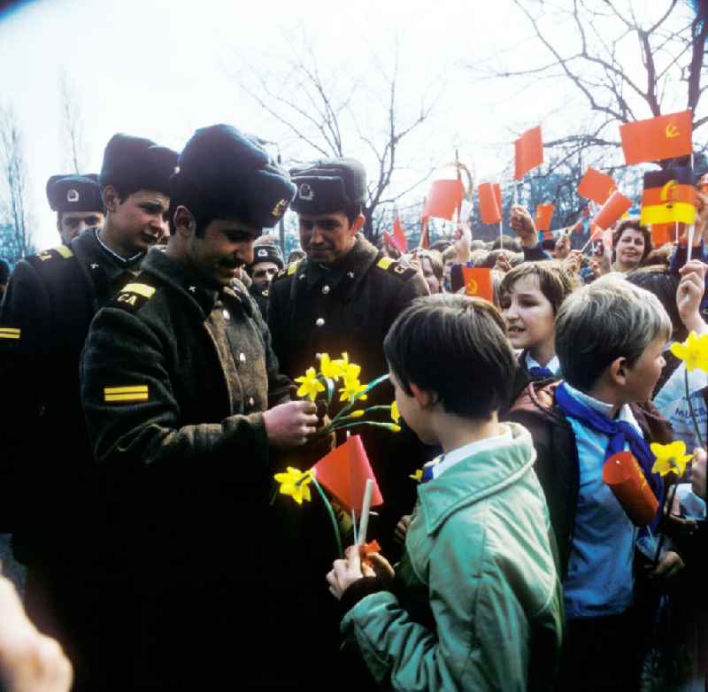 Pioniere / Jungpioniere übergeben Blumen an sowjetische Soldaten anlässlich zum Tag der Befreiung. Schaulustige mit Winkelementen im Hintergrund,  Flagge Sowjetunion und DDR.