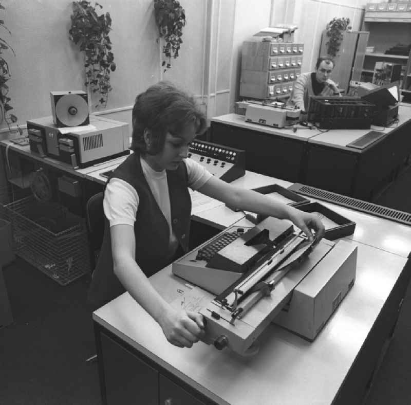 Eine Frau bei der Arbeit vor einer Schreibmaschine im Büro. Sie tippt ein Dokument während ein Kollege zusieht.