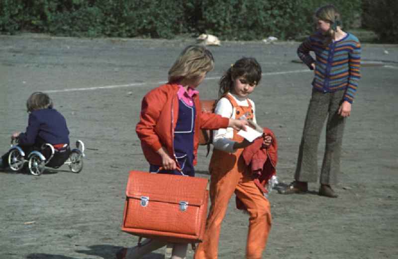 Zwei Mädchen mit Lederranzen gehen nach dem Ende der letzten Unterrichtsstunde über den Sportplatz einer Berliner Schule nach Hause. Obwohl es in der DDR eine gut ausgebaute Hortbetreuung gab, durften viele Schüler bereits in den ersten Klassen sogenannte Schlüsselkinder sein. Sie hatten von ihren Eltern die Erlaubnis, nach der Schule allein nach Hause zu gehen und verbrachten ihre Freizeit oft mit Freunden. Die Eltern waren meist beide berufstätig und kamen erst zum Abendbrot nach Hause.