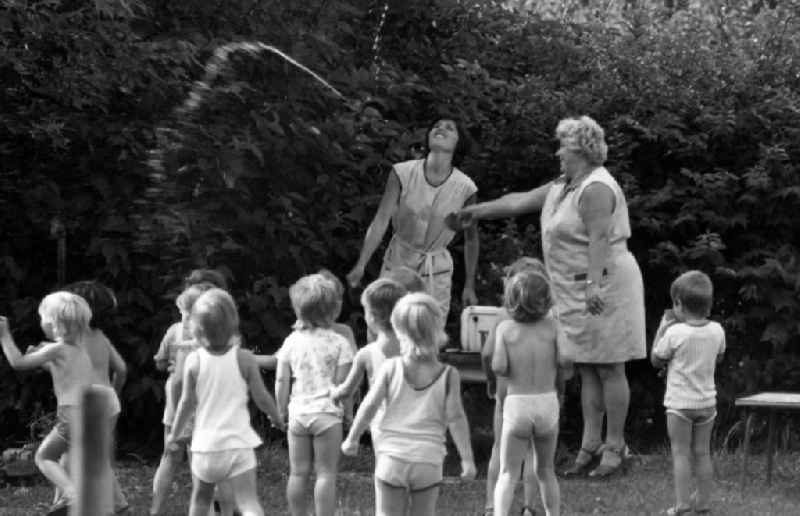 Children in the garden of the kindergarten in summer in Berlin Eastberlin on the territory of the former GDR, German Democratic Republic. Nursery nurse spray water into the air for refreshment