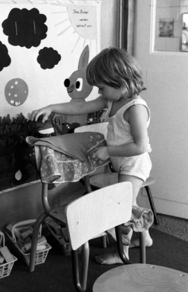 Preparing for a nap in the kindergarten in Berlin Eastberlin on the territory of the former GDR, German Democratic Republic. Toddler tries to undress on his own and pulls on his sock or stocking. Toddler undresses and puts a T-shirt over a chair
