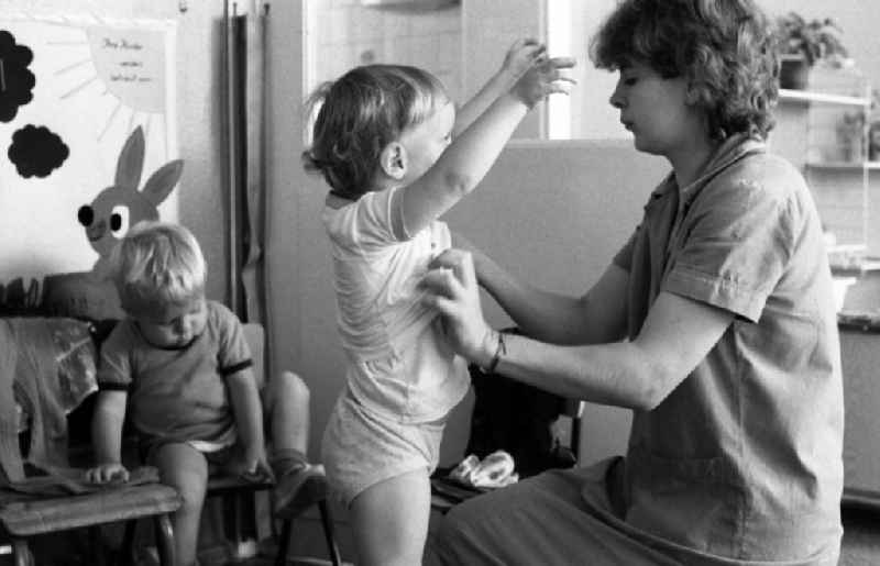 Preparing for a nap in the kindergarten in Berlin Eastberlin on the territory of the former GDR, German Democratic Republic. Nursery nurse helping a toddler undress