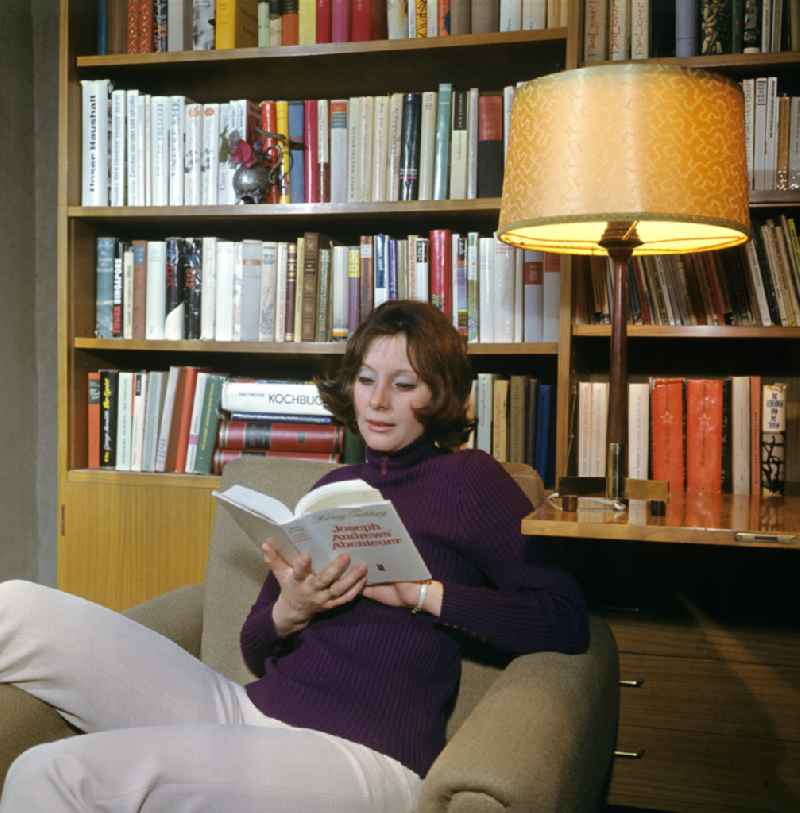 A young woman sits in an armchair and reads a book in front of a bookshelf in a typically furnished living room in the Mitte district of Berlin East Berlin in the territory of the former GDR, German Democratic Republic