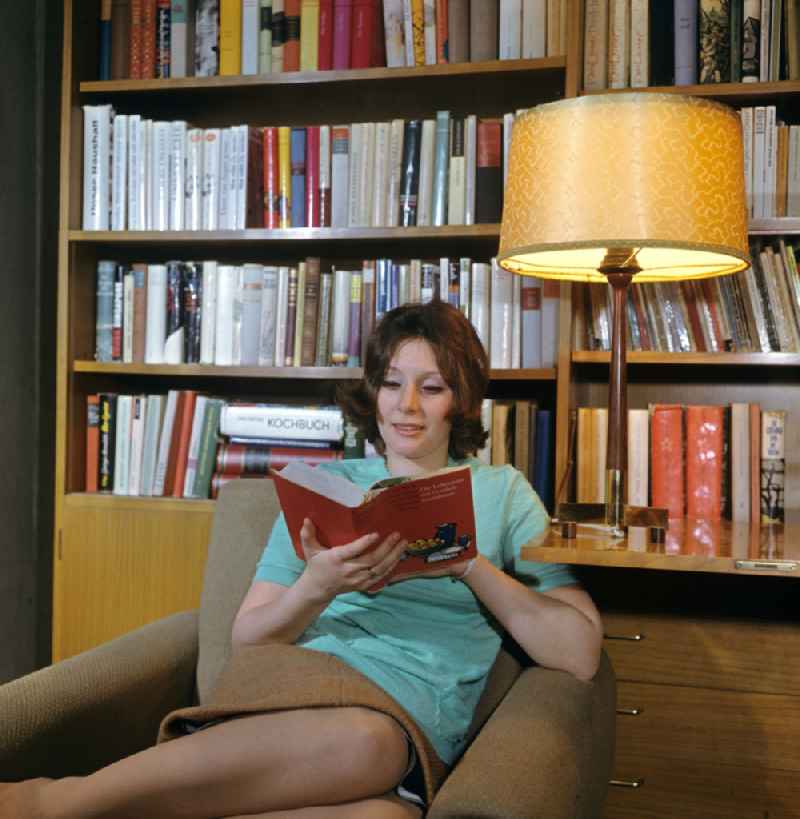 A young woman sits in an armchair and reads a book in front of a bookshelf in a typically furnished living room in the Mitte district of Berlin East Berlin in the territory of the former GDR, German Democratic Republic