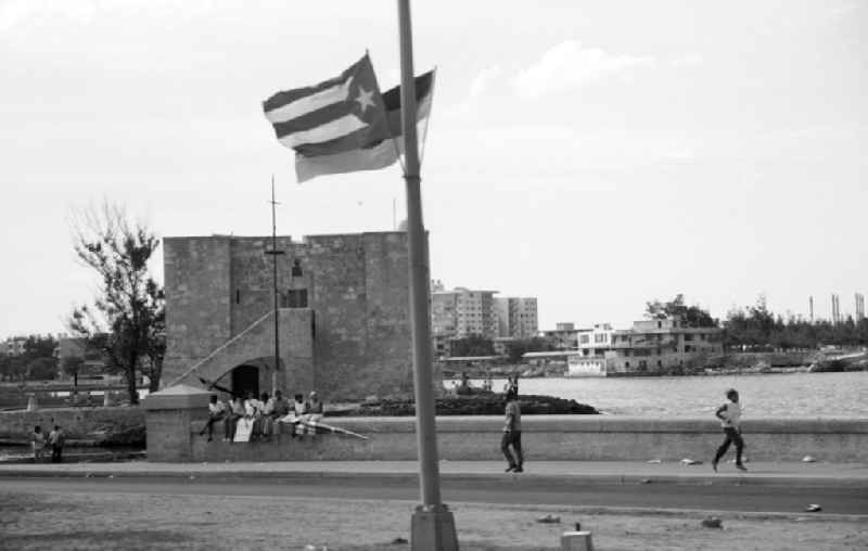 Eine Gruppe Jugendlicher sitzt mit Plakaten zur Ankunft des Staats- und Parteivorsitzenden der DDR, Erich Honecker, auf der Ufermauer an der Promenade Malecón in Havanna. Honecker stattete vom 2