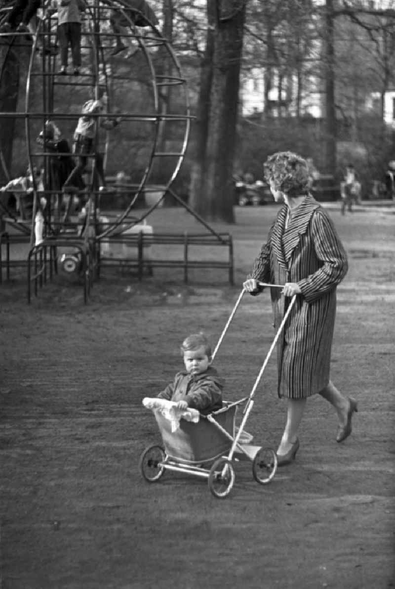 Eine Frau geht mit ihrem Kind in einem Sport-Kinderwagen Marke Eigenbau im Clara-Zetkin-Park in Leipzig spazieren, im Hintergrund spielen Kinder auf einem Klettergerüst.