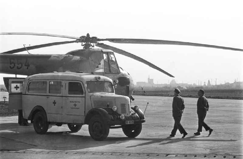 Im Vordergrund zu sehen ist ein ein Krankenwagen vom Typ Robur. Hinter dem Krankenwagen vom Roten Kreuz steht ein Hubschrauber des Typs Mil Mi-4. Die Mil Mi-4 ist ein sowjetischer / russischer Hubschrauber / Helikopter, der von der NVA genutzt wurde. Neben den Fahrzeugen ein Mann in zivil und ein Militär. Bestmögliche Qualität nach Vorlage!