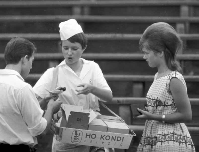 Eine Verkäuferin der HO Kondi - mit Bauchladen - verkauft Süßigkeiten auf dem Turnfest. Neben ihr ein Mädchen mit Toupet-Frisur und einem Kleid im Stil der 196