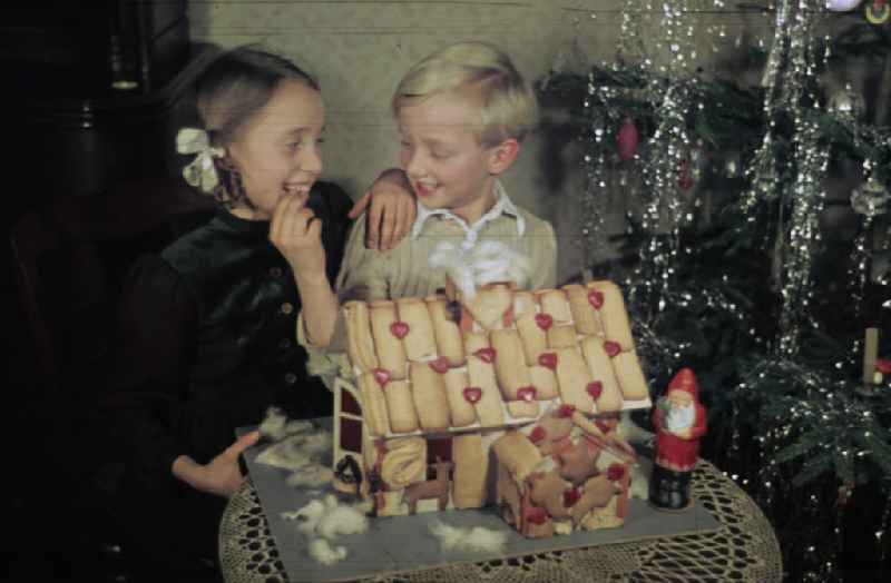 Weihnachten in der Nachkriegszeit. Geschwister knabbern am Lebkuchenhaus vor dem Weihnachtsbaum an Heiligabend. Christmas in the postwar period. Brothers and sisters / sibs nibble on a gingerbread house in front of the Christmas tree at the Christmas Eve.