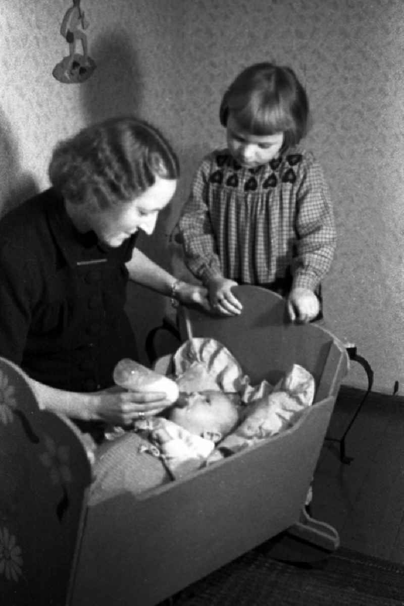 A mother gives the teat bottle to her baby in the cradle and the big sister looks to in Merseburg in the federal state Saxony-Anhalt in the area of the former GDR, German democratic republic