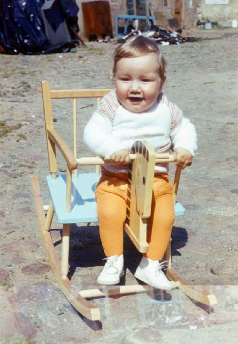 A small child with yellow sock trousers sits laughing in a wood rocking horse in Neustrelitz in the federal state Mecklenburg-West Pomerania in the area of the former GDR, German democratic republic
