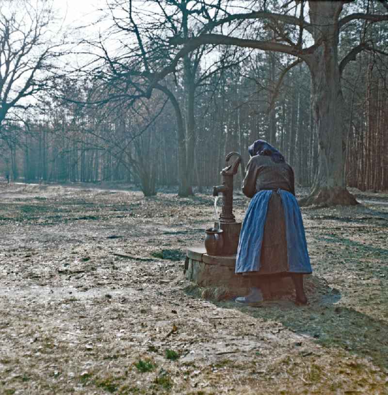 Scene from the film and television production 'Rublak - The Legend of the Surveyed Land' in Heide, Saxony, in the area of the former GDR, German Democratic Republic. An old farmer's wife in everyday dress stands at the edge of a forest at a hand pump. Water flows into a clay jug. The actress Doris Thalmer as a Sorbian farmer's wife