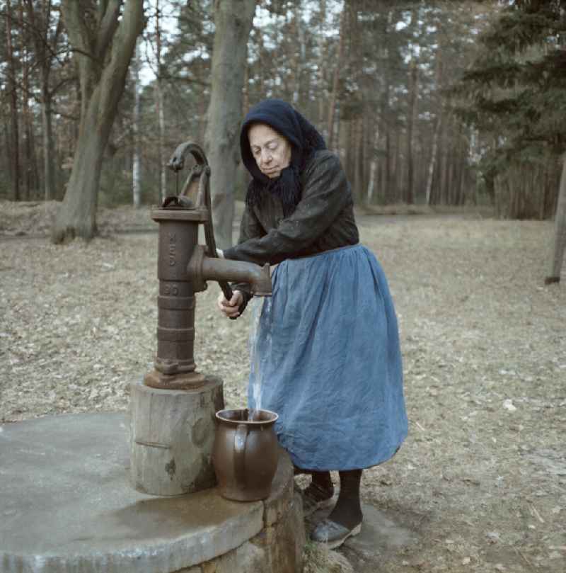 Scene from the film and television production 'Rublak - The Legend of the Surveyed Land' in Heide, Saxony, in the area of the former GDR, German Democratic Republic. An old farmer's wife in everyday dress stands at the edge of a forest at a hand pump. Water flows into a clay jug. The actress Doris Thalmer as a Sorbian farmer's wife