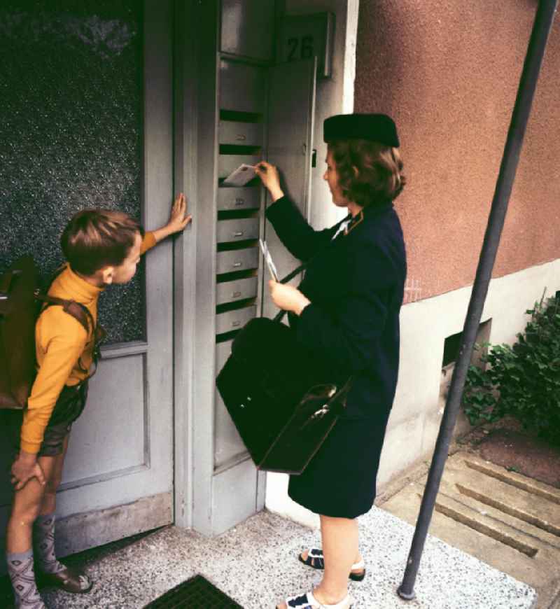 Briefträgerin beim Einwerfen der Post in Briefkästen eines Wohnhauses. Ein Junge guckt zu.