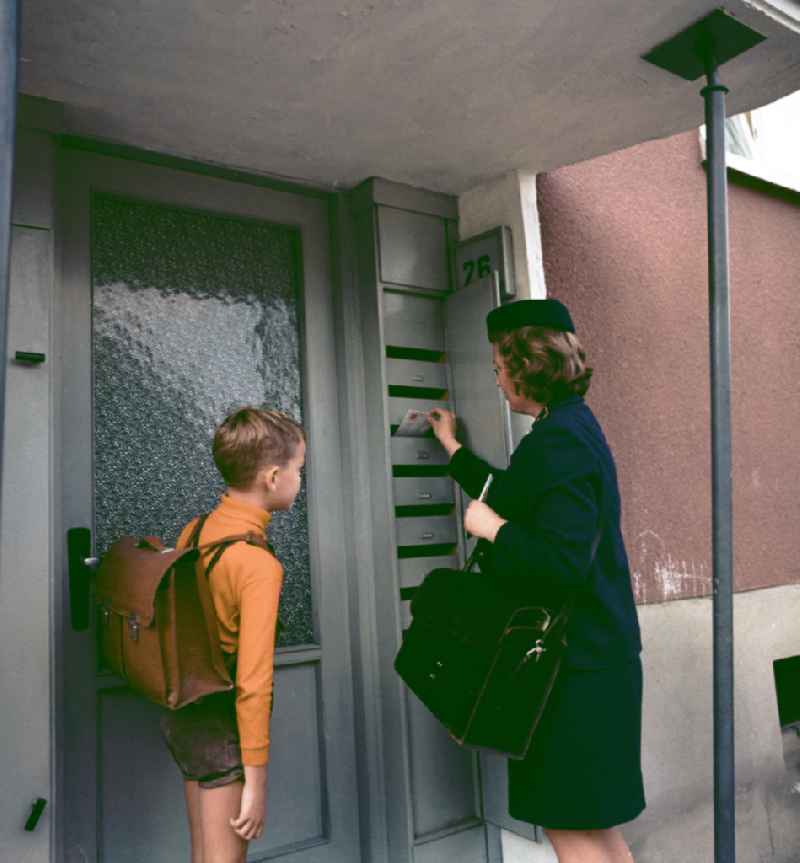 Briefträgerin beim Einwerfen der Post in Briefkästen eines Wohnhauses. Ein Junge guckt zu.