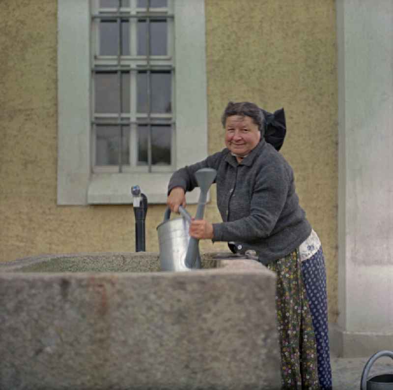 Scene from the film and television production 'Portrait of a Center' on the main street in Ralbitz-Rosenthal, Saxony in the area of the former GDR, German Democratic Republic. A brick water basin. A Sorbian woman in everyday dress lifts a metal watering can out of the water and looks into the camera