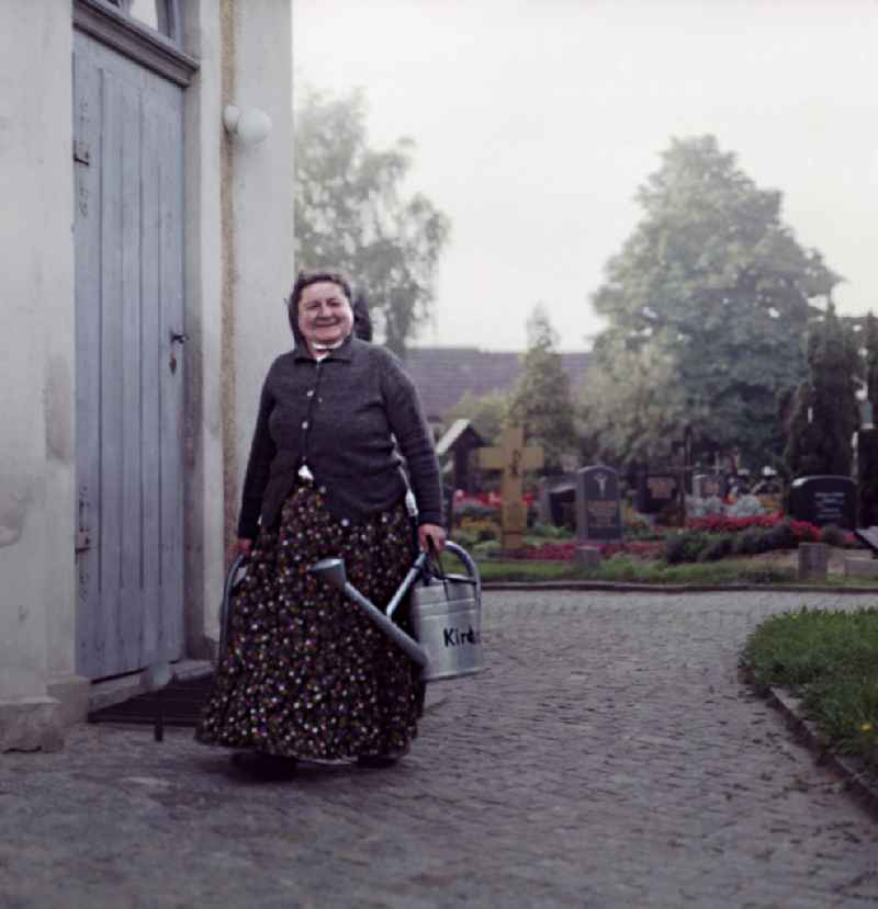 A Sorbian woman in everyday costume carries a metal watering can as a scene from the film and television production ' Portrait of a Center ' Sorbian cemetery in Ralbitz-Rosenthal, Saxony in the area of the former GDR, German Democratic Republic