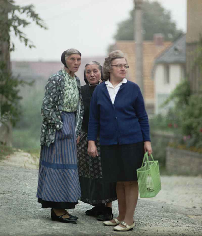 Three women stand on a village street. Two older women wear Sorbian everyday costume as a scene from the film and television production 'Portrait of a Center' on the main street in Raeckelwitz, Saxony in the area of the former GDR, German Democratic Republic