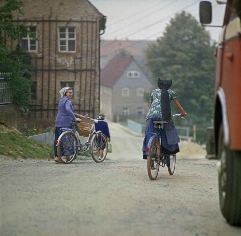 Two female cyclists in traditional peasant clothing meet on a village street as a scene from the film and television production 'Portrait of a Center' on the main street in Raeckelwitz, Saxony in the area of the former GDR, German Democratic Republic