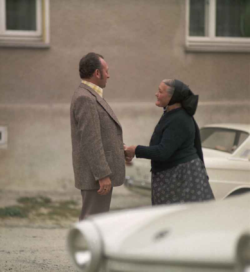 On a village street, a man and an elderly woman in Sorbian everyday costume stand between two Trabant cars and have an animated conversation as a scene from the film and television production 'Portrait of a Center' on the main street in Raeckelwitz, Saxony in the area of the former GDR, German Democratic Republic