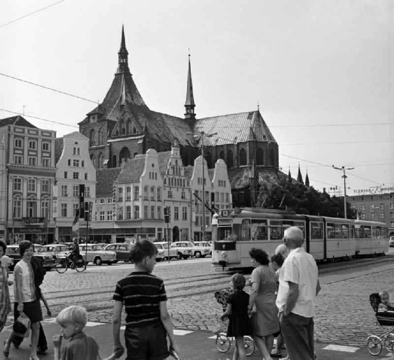 Blick über den Neuen Markt mit Straßenbahn, den bekannten Giebelhäusern und der St.-Marien-Kirche. Menschen überqueren die Strasse. In Rostock wird gerade die Ostseewoche zelebriert, eine jährlich in der DDR veranstaltete internationale Festwoche. Veranstalter dieser zwischen 1958 und 1975 stattfindenden Festwoche war der FDGB (Freier Deutscher Gewerkschaftsbund). Zahlreiche politische, kulturelle und sportliche Veranstaltungen fanden zu dieser Zeit statt. Das Motto der Veranstaltung lautete: 'Die Ostsee muss ein Meer des Friedens sein'.