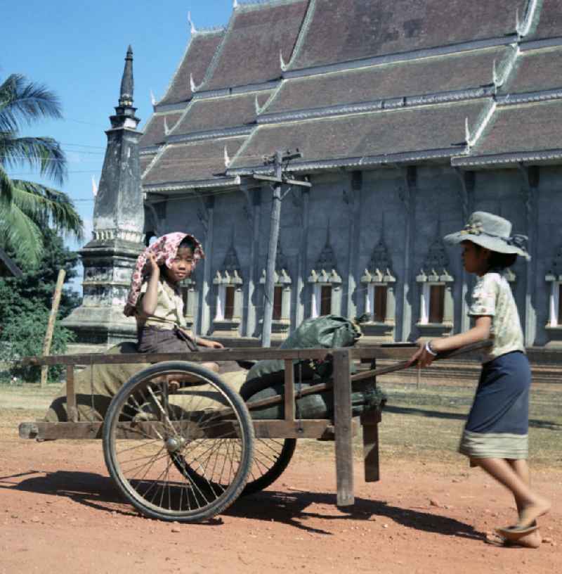 Kinder mit Karren vor einem zur goldenen Stupa Pha That Luang gehörenden Tempel in Vientiane, der Hauptstadt der Demokratischen Volksrepublik Laos.