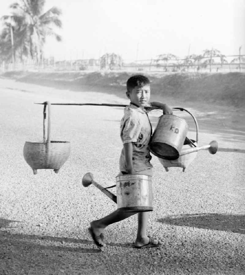 Ein Junge trägt Gießkannen und geschulterte Körbe auf einer Straße in Vientiane, der Hauptstadt der Demokratischen Volksrepublik Laos.