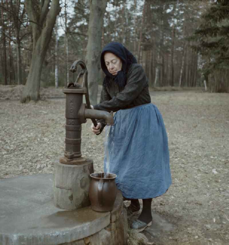 Scene from the film and television production 'Rublak - The Legend of the Surveyed Land' in the Haide district of Weisskeissel, Saxony in the area of the former GDR, German Democratic Republic. At the edge of the forest, a farmers wife in Sorbian work costume stands at a water pump and fills water into a clay jug