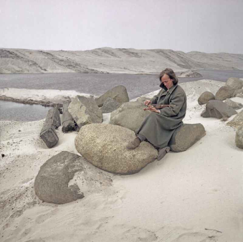 Scene from the film and television production 'Rublak - The Legend of the Surveyed Land' in Weisswasser/Oberlausitz, Saxony in the area of the former GDR, German Democratic Republic. The actor Christian Grashof sits playing the violin on a very large boulder in the middle of a sandy plain in a waste landscape of a brown coal open-cast mine